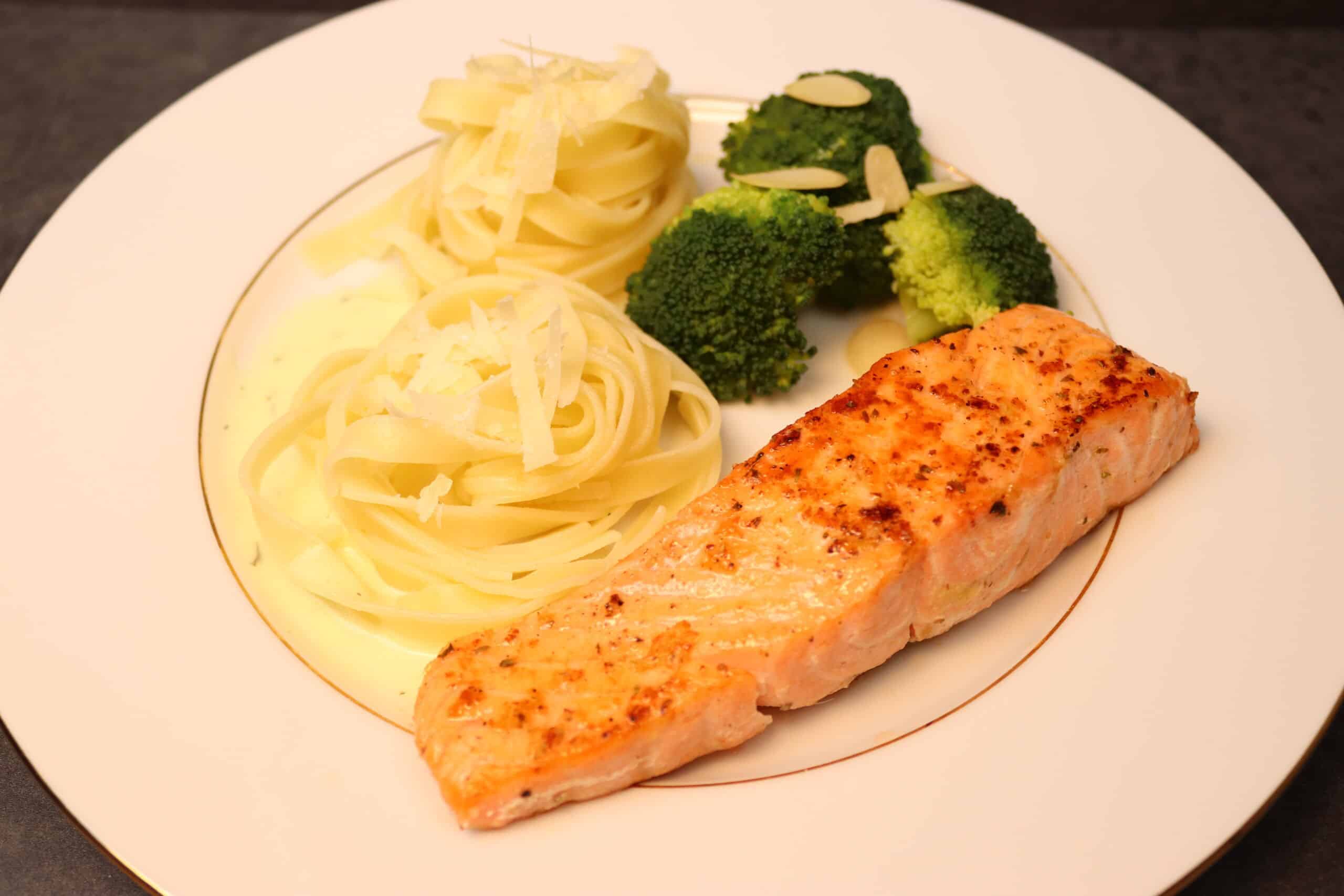 Ein leckeres gold-gelb gebratenes Lachsfilet mit Bandnudeln mit Käse und Brokkoli mit Mandelblättern auf einem Teller serviert.